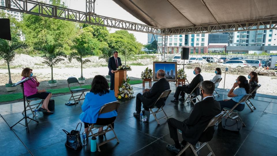 A man wearing a suit and tie and speaking from a pulpit outside, with around eight people wearing face masks and sitting in chairs socially distanced.