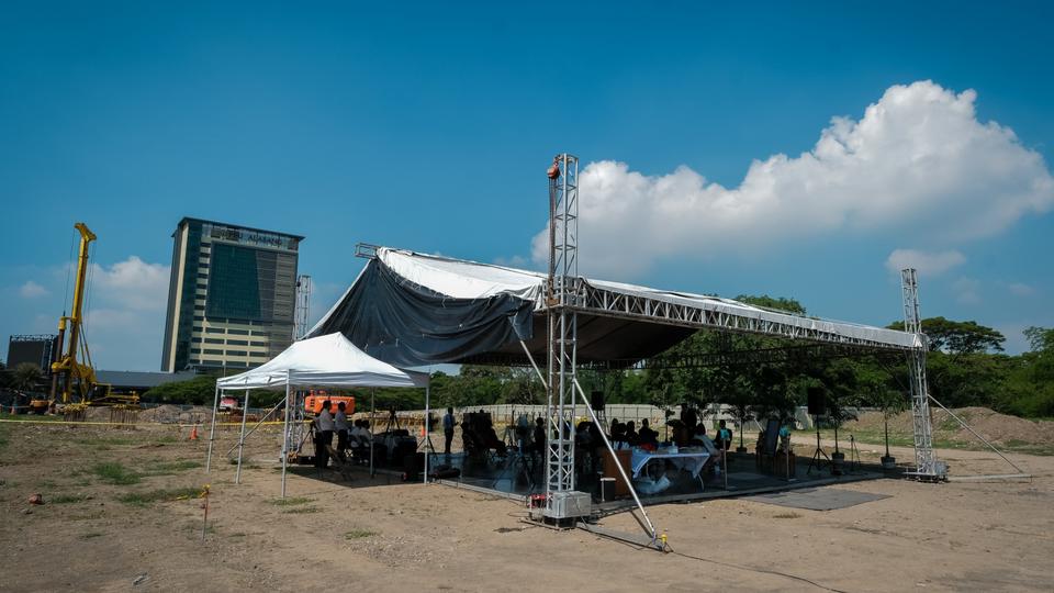 A congregation of participants in a dirt field and under a large cloth tent, with trees and a tall building in the background.