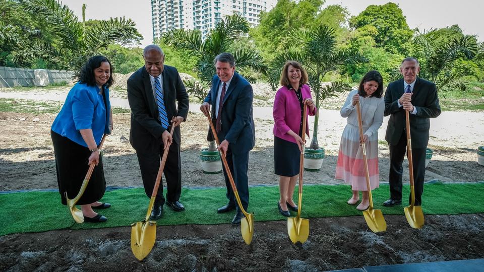 A row of three men in suits and three women in dresses holding ceremonial golden shovels into the ground.