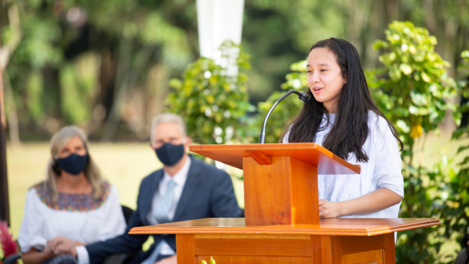 A young woman speaking from a pulpit outside, with two people sitting in chairs in the background.