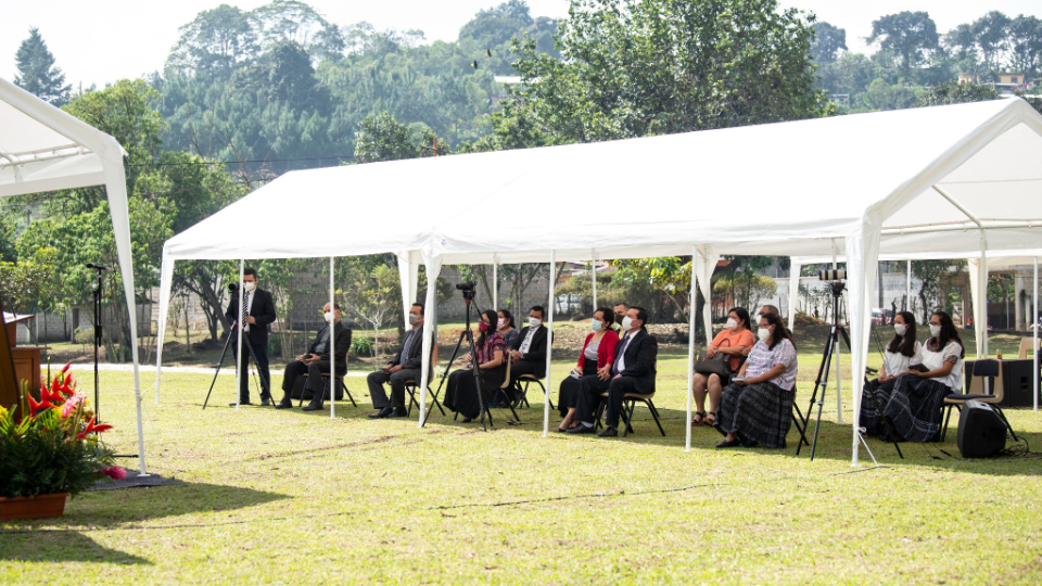 A group of around 10 people sitting in chairs outside under a cloth shade structure.