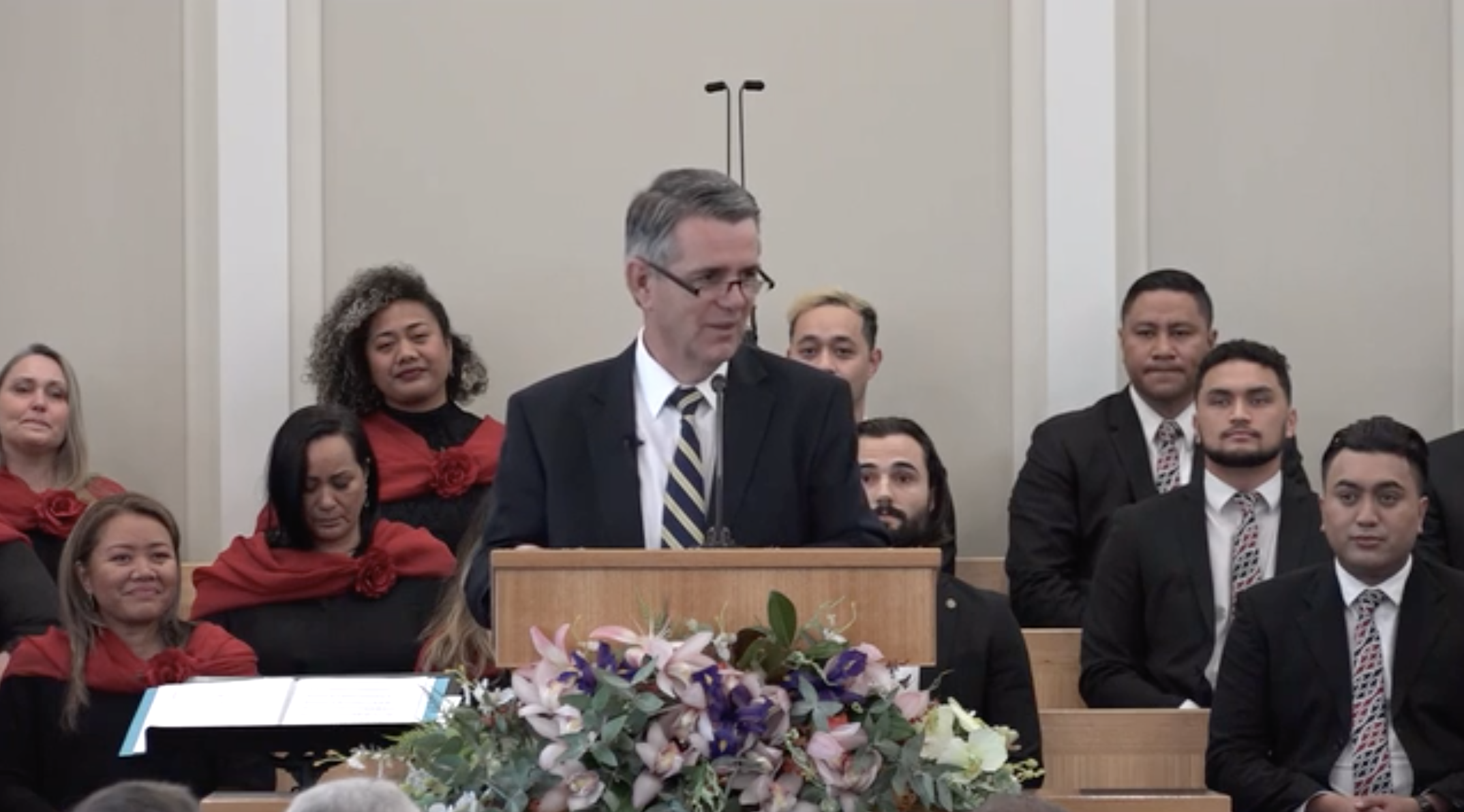 A man wearing a suit, tie and glasses and speaking from a pulpit outside.